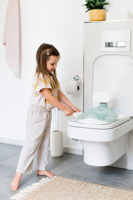 Mergaitė vonios kambaryje laiko šviesiai žalią banginio formos tualeto dangtį, uždėtą ant klozeto sėdynės