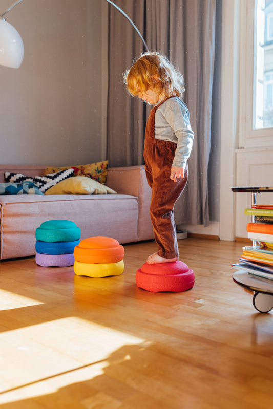 Vaikas žaidžia kambaryje stovėdamas ant raudono Stapelstein akmenuko, šalia sukrauti spalvoti akmenukai žaidimams ir balansavimui